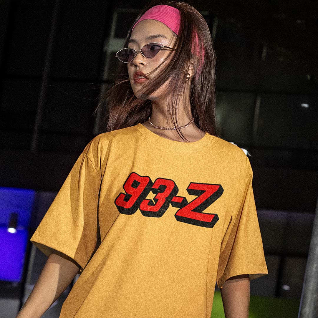 A woman wearing a yellow oversized t-shirt with a red and black 3D-effect '93-Z' graphic on the chest, paired with a pink headband and glasses, set against a dark, moody background.