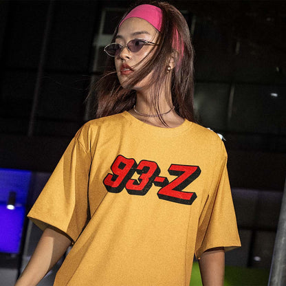 A woman wearing a yellow oversized t-shirt with a red and black 3D-effect '93-Z' graphic on the chest, paired with a pink headband and glasses, set against a dark, moody background.
