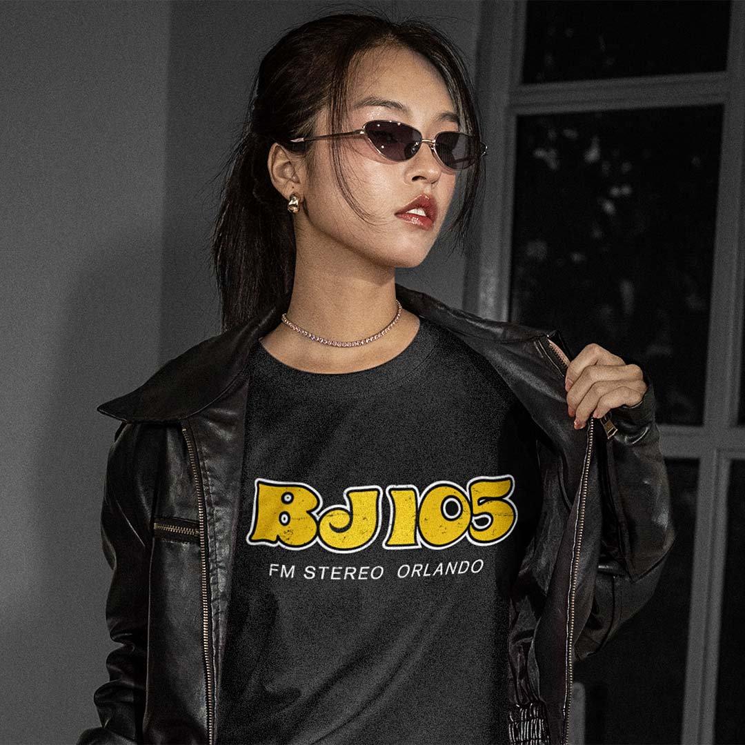A woman wearing a black leather jacket over a black graphic t-shirt with yellow and white 'BJ105 FM STEREO ORLANDO' text, styled with dark sunglasses and a delicate necklace in a dimly lit indoor setting.