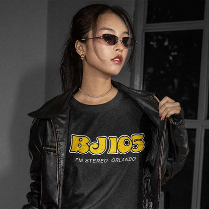 A woman wearing a black leather jacket over a black graphic t-shirt with yellow and white 'BJ105 FM STEREO ORLANDO' text, styled with dark sunglasses and a delicate necklace in a dimly lit indoor setting.