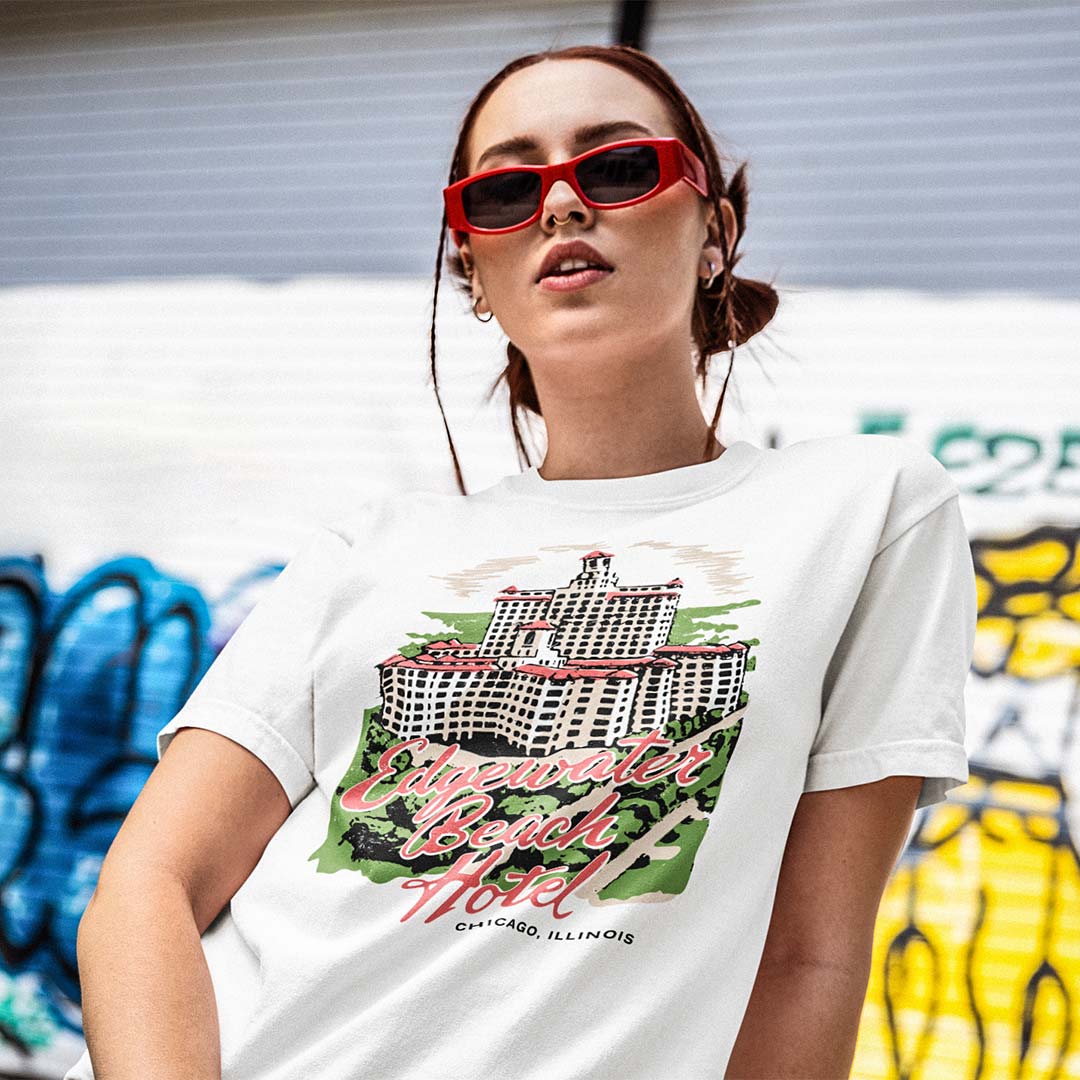 A woman wearing red sunglasses and a white graphic t-shirt with a colorful illustration of the Luxor Hotel in Chicago, set against a blurred graffiti wall background.