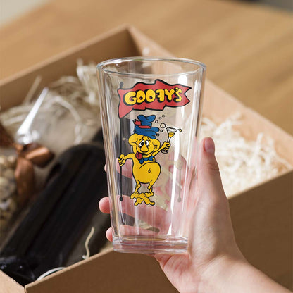 A hand holds a clear glass pint tumbler featuring a colorful 'Goofy's' cartoon character print, with a cardboard shipping box and shredded paper visible in the background.