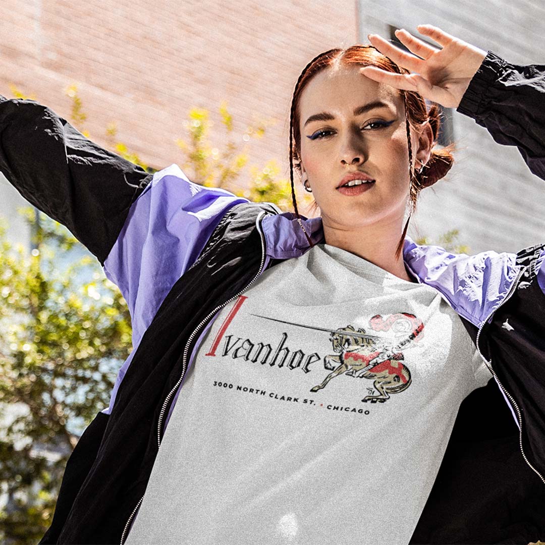 A woman wearing a white graphic t-shirt with 'Ivanhoe' and a lion illustration, layered under a black and purple windbreaker jacket, photographed outdoors with a blurred brick wall and greenery in the background.