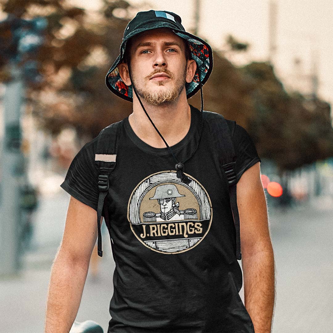 A man wearing a black t-shirt with a circular J. Rigging graphic, a bucket hat, and a backpack, standing outdoors on a blurred street background.