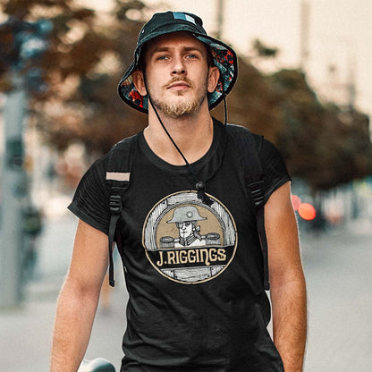 A man wearing a black t-shirt with a circular J. Rigging graphic, a bucket hat, and a backpack, standing outdoors on a blurred street background.