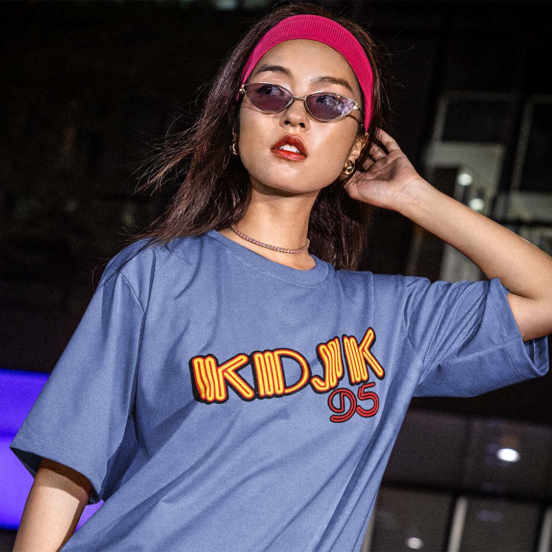 A woman wearing a blue oversized graphic T-shirt with 'KDJK 95' printed across the chest in orange and red, styled with a pink headband and sunglasses in a dark nighttime setting.