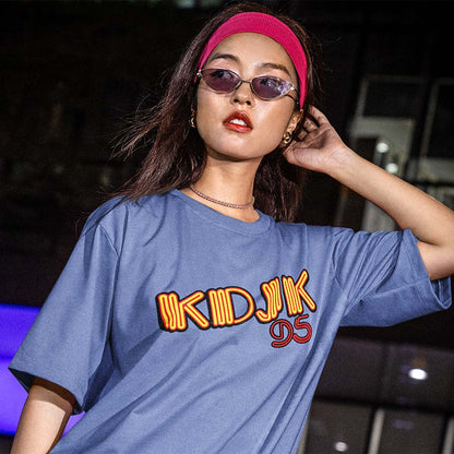 A woman wearing a blue oversized graphic T-shirt with 'KDJK 95' printed across the chest in orange and red, styled with a pink headband and sunglasses in a dark nighttime setting.