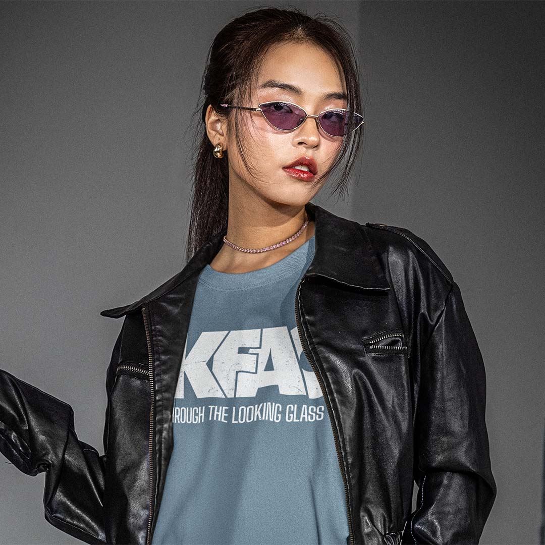 A woman wearing a black leather jacket over a slate blue graphic t-shirt with white text, styled with purple-tinted sunglasses and a delicate silver necklace, set against a plain gray studio background.