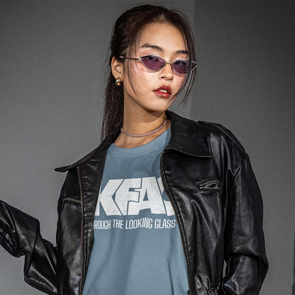 A woman wearing a black leather jacket over a slate blue graphic t-shirt with white text, styled with purple-tinted sunglasses and a delicate silver necklace, set against a plain gray studio background.