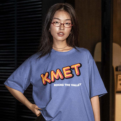 A woman wearing a blue oversized graphic T-shirt with 'KMET' in red and yellow letters and 'ROCKS THE VALLEY' in white, posed against a dark indoor background.