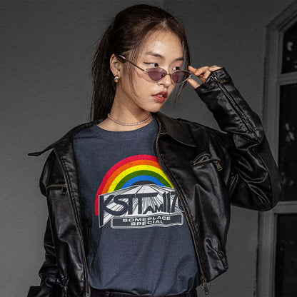 A woman wearing a black faux-leather moto jacket over a dark gray graphic t-shirt with a rainbow and 'KSTT AM 1170' print, styled with sunglasses against a dark gray background.