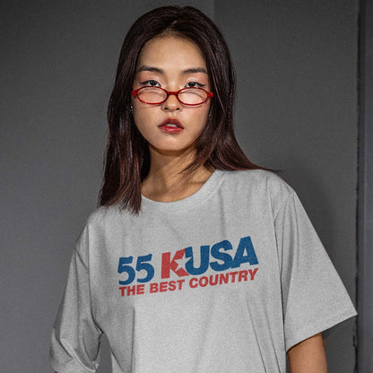 A model wearing a light gray oversized t-shirt with a red and blue '55 KUSA THE BEST COUNTRY' graphic on the front, paired with red-framed glasses, set against a dark gray studio background.