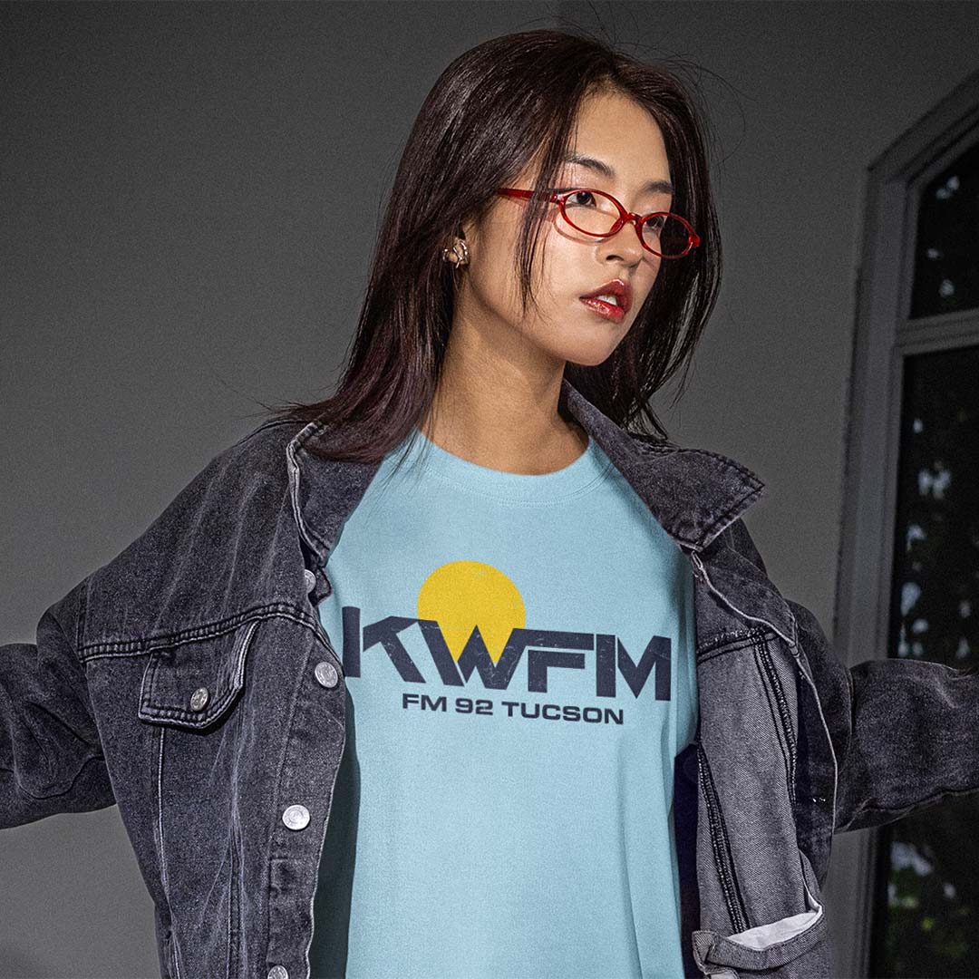 A woman wearing a light blue graphic t-shirt with a yellow sun and black 'KWFM FM 92 TUCSON' print, layered under a faded black denim jacket. She is wearing red-framed glasses and looking to the side against a dark gray background.