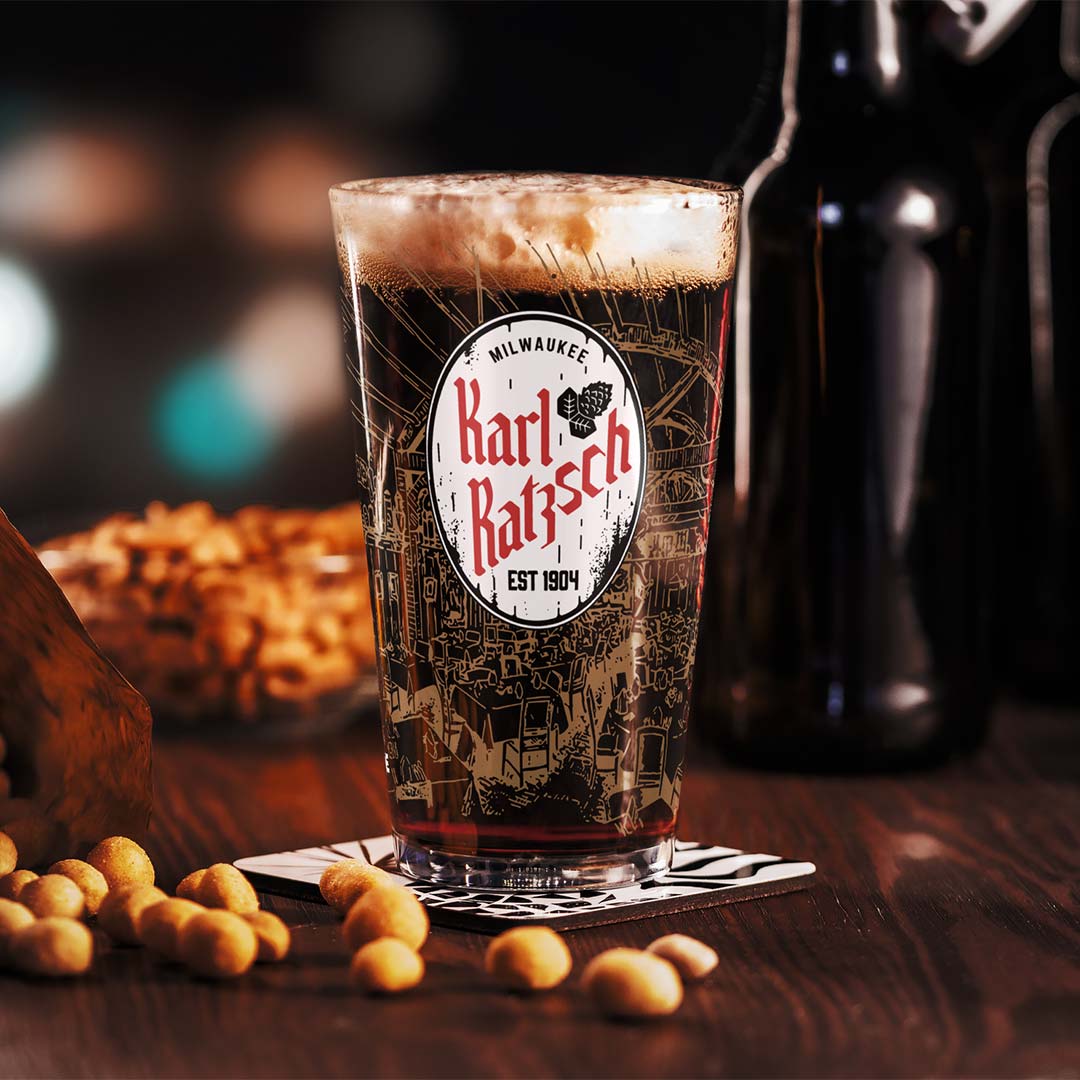 A clear pint glass with a printed Milwaukee Karl Batsch logo, filled with dark beer and topped with foam, set on a coaster on a wooden table with peanuts and a dark bottle in the background.