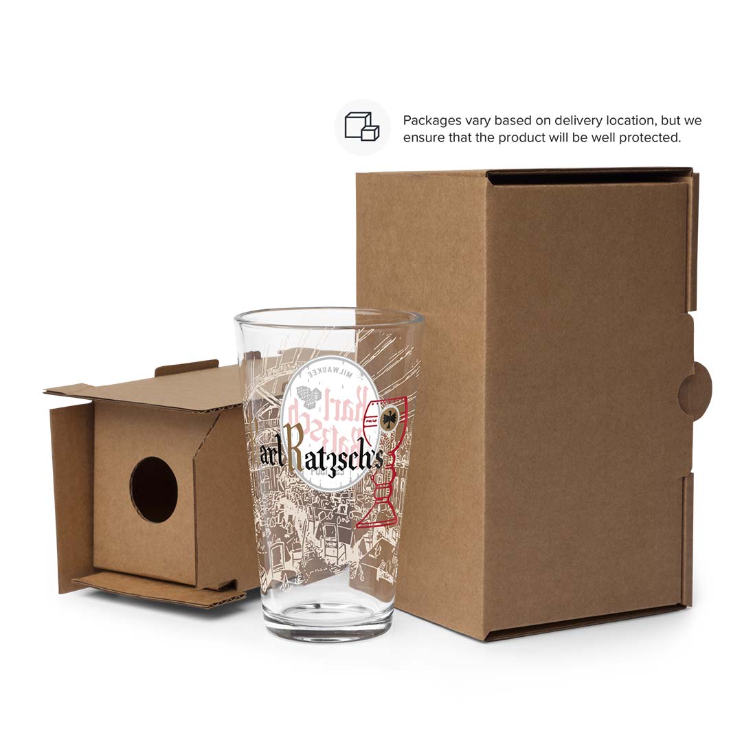 A clear glass tumbler with printed artwork and the text 'and Ratschke' stands in front of a brown cardboard box, with the box lid placed beside it on a white background. A small text note at the top says packaging varies based on delivery location, but the product is well protected.