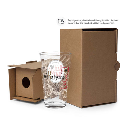 A clear glass tumbler with printed artwork and the text 'and Ratschke' stands in front of a brown cardboard box, with the box lid placed beside it on a white background. A small text note at the top says packaging varies based on delivery location, but the product is well protected.