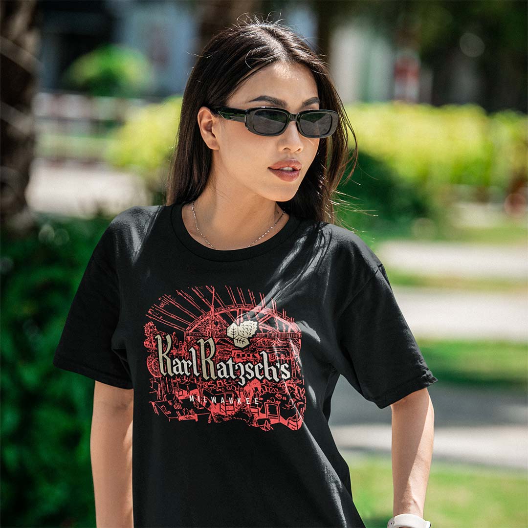 A woman wearing a black Karl Ransom's t-shirt with a red and white graphic print, photographed outdoors in a sunny park setting.