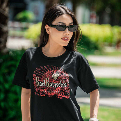 A woman wearing a black Karl Ransom's t-shirt with a red and white graphic print, photographed outdoors in a sunny park setting.