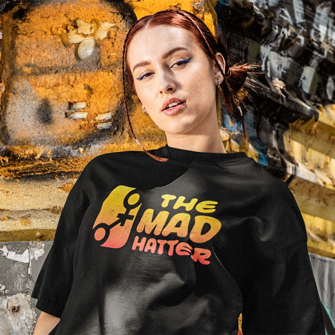 A woman with red hair wearing a black t-shirt with a colorful graphic that reads 'THE MAD HATTER' in yellow, orange, and red, posed against a textured, weathered yellow and gray wall.