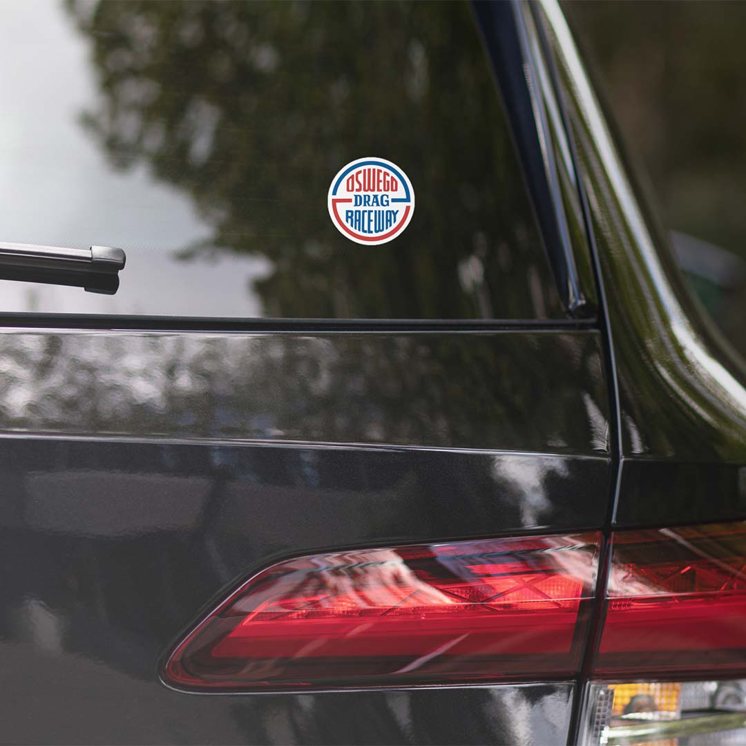 A circular 'Oswego Drag Racing' decal applied to the rear window of a black car, with reflections of trees visible on the glass.
