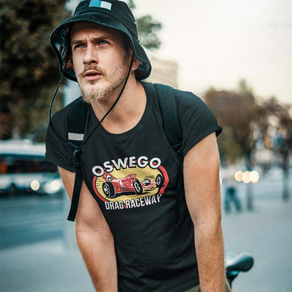 A man wearing a black graphic t-shirt with a red and yellow race car design and the text 'OSWEGO DRAG-RACEWAY', along with a dark bucket hat and a backpack, set against a blurred city street background.