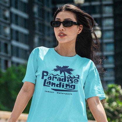 A woman wearing a light blue graphic t-shirt with a dark navy palm tree and 'Paradise Landing' text, photographed outdoors against a blurred background of buildings and greenery.