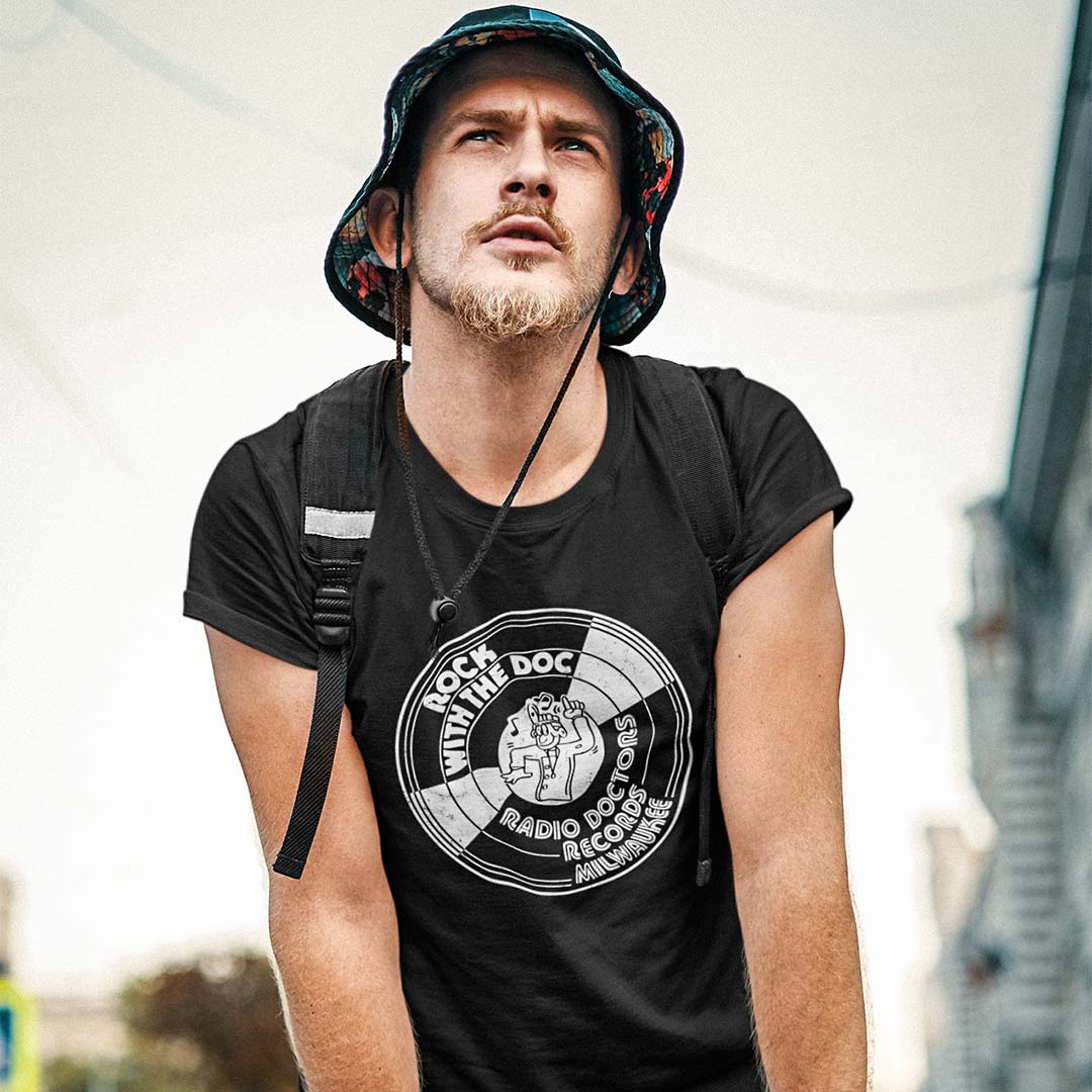 A man wearing a black graphic t-shirt with a white circular 'With The Doc Radio Docs Records' print, styled with a patterned bucket hat and a black backpack strap over his shoulder.