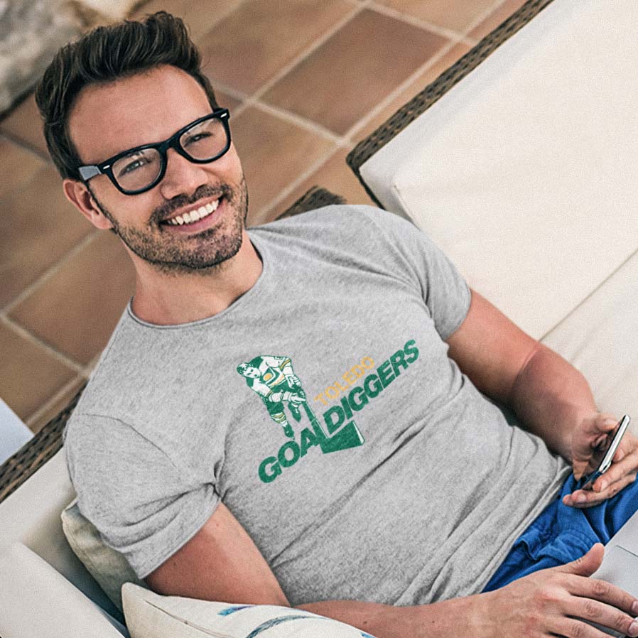 A smiling man wearing black-rimmed glasses and a heather gray t-shirt with a green and yellow 'GOAL DIGGERS' graphic, reclining on a white patio sofa.