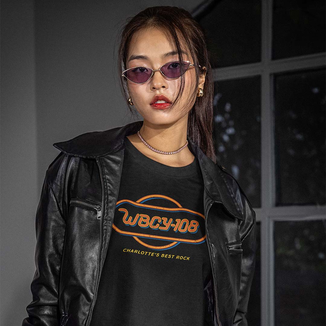 A woman wearing a black leather jacket and a black graphic t-shirt with orange and yellow text reading 'WBCY108' and 'CHARLOTTE'S BEST ROCK', posed in front of a dark windowed wall.