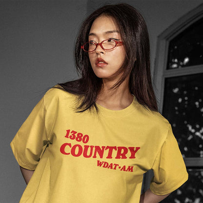 A woman with dark hair and red-framed glasses wearing an oversized yellow t-shirt with red text reading '1380 COUNTRY WDATAM'.