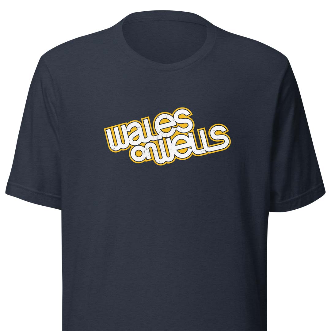 A navy blue short-sleeve t-shirt with a retro-style graphic on the chest reading 'wales on wells' in white bubble letters with a yellow outline.
