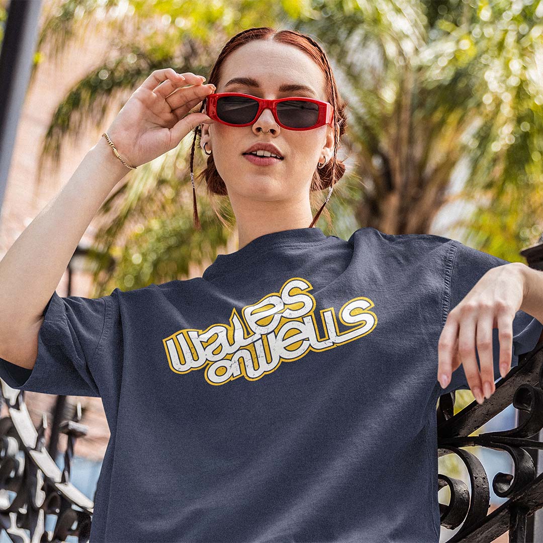 A woman outdoors wearing a navy blue graphic t-shirt with 'waves on wells' printed in yellow and white across the chest, and red-framed sunglasses.