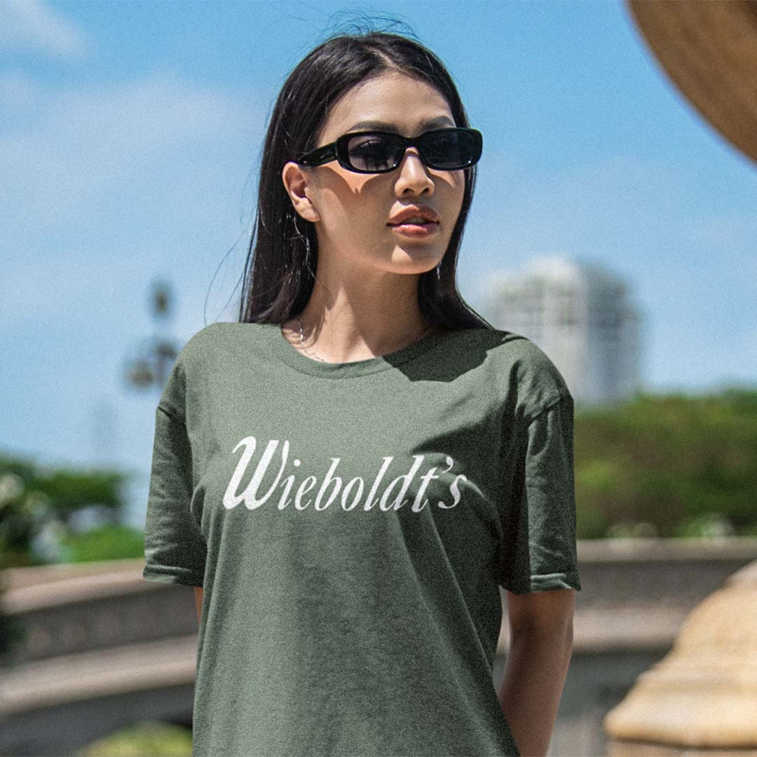 A woman wearing a dark olive green t-shirt with 'Wieboldt's' printed in white script across the chest, photographed outdoors against a blurred background of blue sky and greenery.