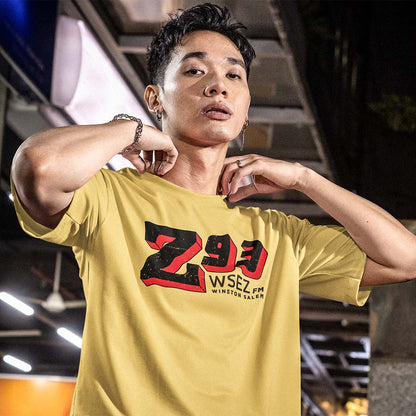 A man wearing a yellow t-shirt with a bold black and red graphic on the chest that reads 'Z93 WSEZ WINSTON SALEM', posing in a dimly lit indoor environment.