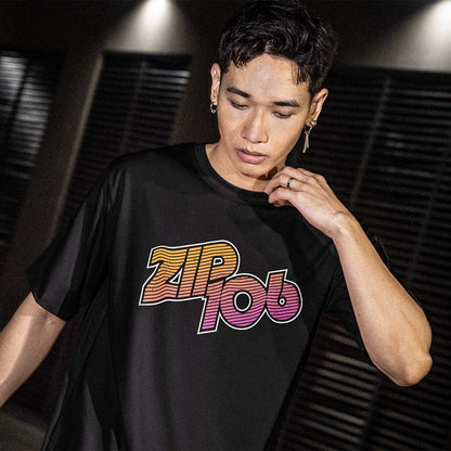 A man wearing a black t-shirt with a colorful 'ZIP 106' graphic on the chest, posed against a dark background with horizontal blinds.